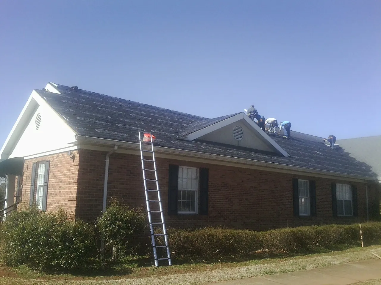 Crew tearing shingles off a brick ranch roof with a ladder set — Emergency Roof Repair work by our crew in East Meadow