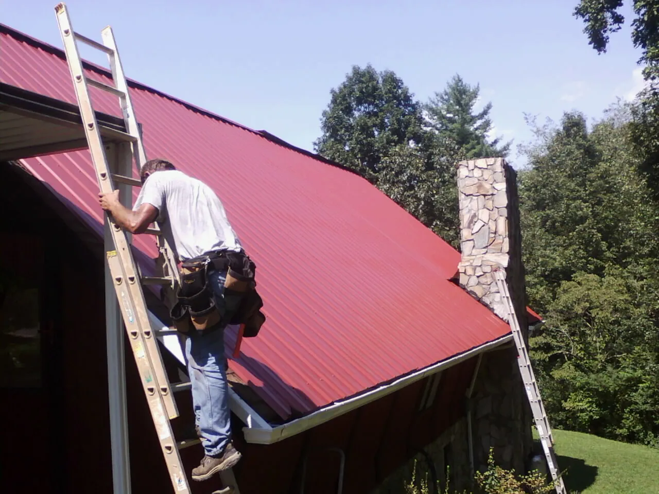 Roofer on a ladder beside a red metal roof and stone chimney — Soffit & Fascia Repair by Roofing Brothers in East Meadow